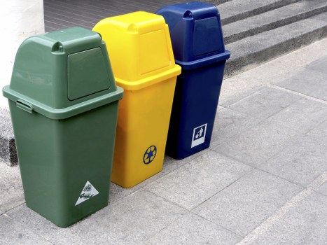 Three upright waste bins placed on a paved sidewalk surface, positioned in a row from left to right. The first bin on the left is green with a slightly textured surface and a triangular recycling symbol on the front lower section. The middle bin is bright yellow with a smooth finish and a small circular symbol near the bottom. The third bin on the right is dark blue with a smooth surface, featuring a white recycling box icon near the bottom. The bins are set against a grey concrete or stone resurfaced pavement, with the top edges of the bins visible and lids closed. The background shows a curb or step edge, indicating they are likely located on a street or pavement area suitable for rubbish disposal and collection services such as those provided by Garden Clearance Plumstead. The overall scene is evenly lit, likely outdoors during daytime, capturing typical waste management equipment for local rubbish collection in Plumstead, London.
