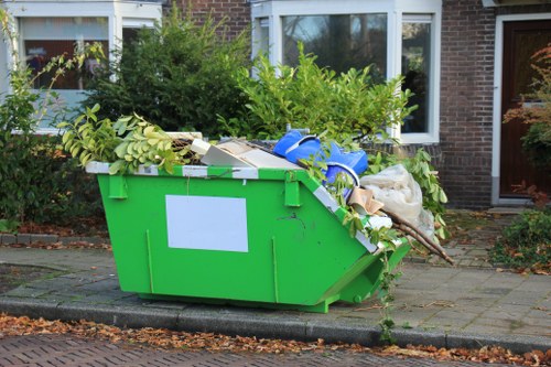 A large, bright green plastic waste container overflowing with garden rubbish is positioned on a paved driveway in front of a residential building. The container is filled with various green plant cuttings, branches, and leaves, with some foliage spilling over the top edges. In addition to plant debris, there are items such as cardboard boxes, plastic wrapping, and miscellaneous refuse contributing to the cluttered appearance. The driveway is bordered by a curb and is adjacent to a brick-fronted house with white-framed windows and a brown door, suggesting a suburban setting. Surrounding the container are some small shrubs and plants, with autumn leaves scattered on the ground. The scene captures a typical garden clearance scenario where Garden Clearance Plumstead might assist with rubbish removal in the local area, emphasizing the type of garden waste collected and disposed of professionally.