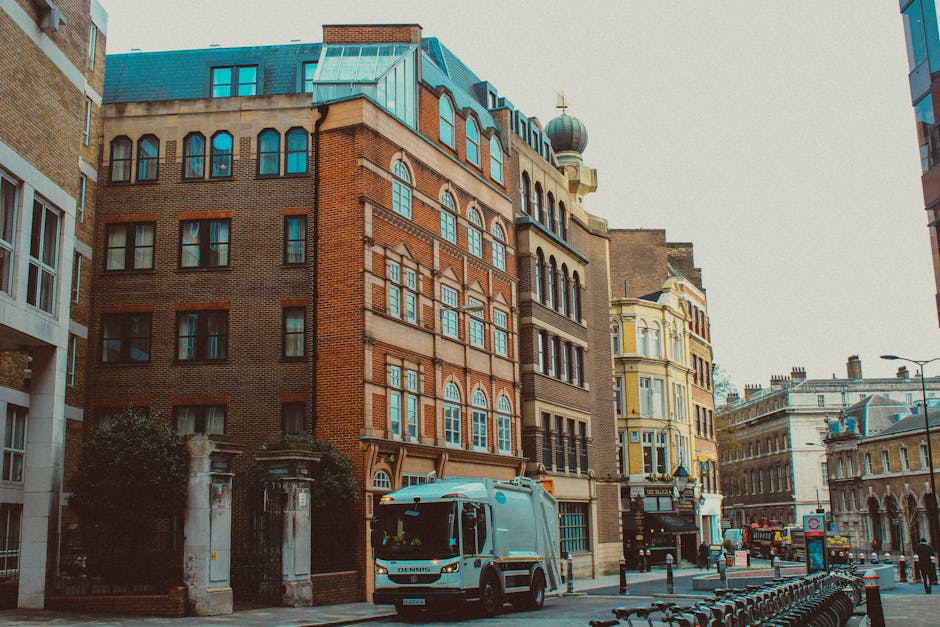 A city street scene featuring several multistory buildings with varied architectural styles, including red brick and ornate facades. In the foreground, a white waste collection vehicle with a compact,