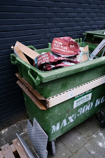 A city street scene featuring several multistory buildings with varied architectural styles, including red brick and ornate facades. In the foreground, a white waste collection vehicle with a compact,