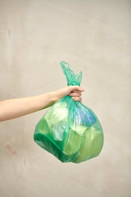 A human arm extending from the left side of the image holds a green plastic refuse bag filled with waste, with the bag's top gathered and tied in a knot. The bag is semi-transparent, showing the conte