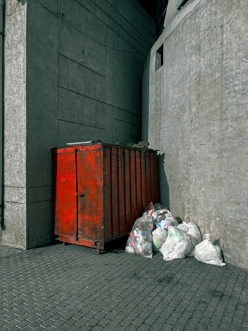 A red metal skip with a weathered, slightly rusted surface is positioned against a dark, textured wall on the left side of the image and a concrete wall on the right side, situated on a paved urban si